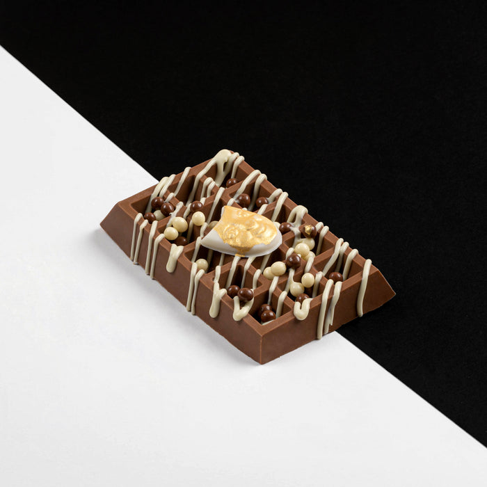 A decorative milk chocolate waffle bar topped with caramel pearls, drizzled white chocolate, and a caramel-filled chocolate shell, displayed on a black and white geometric surface