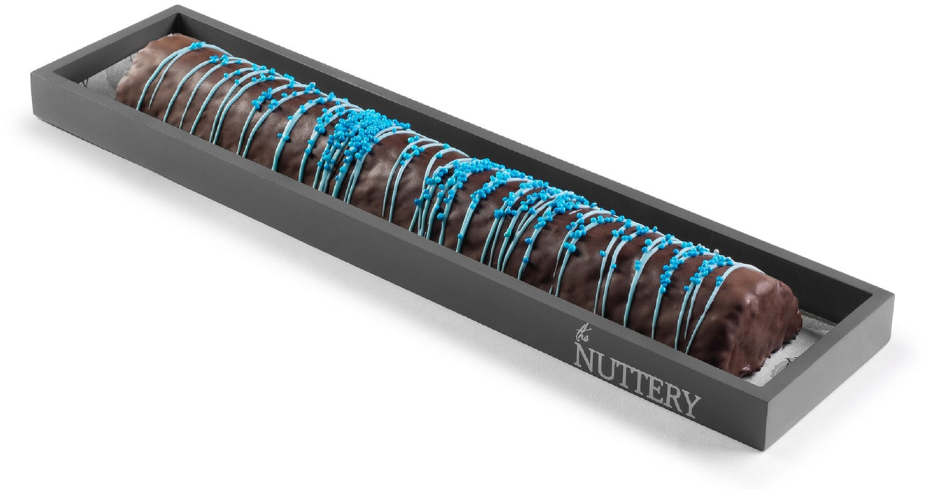A luxurious peanut chew log coated in dark chocolate, topped with bright blue drizzle and sprinkles, displayed in a black Nuttery-branded tray for an eye-catching presentation.