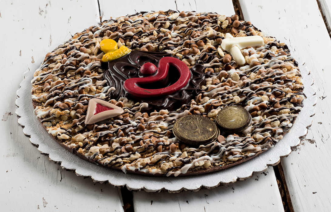 A festive Purim-themed chocolate pie topped with colorful clown decorations, chocolate coins, and hamantaschen-shaped chocolates, drizzled with white and dark chocolate on a scalloped board