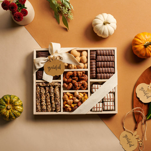 A collection of elegantly arranged wooden gourmet gift trays filled with premium chocolates, roasted nuts, and granola bars. Each tray is wrapped with a neutral fabric ribbon and a custom wooden tag. Set against a modern gray background with scattered chocolates and nuts, this luxurious assortment is perfect for personalized gifting.