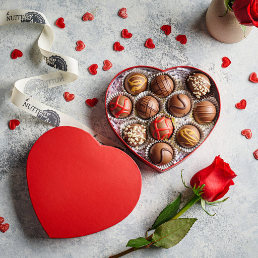 A red heart-shaped gift box filled with assorted gourmet chocolate truffles decorated with caramel drizzle, crushed nuts, and colorful designs, styled alongside a single red rose and scattered heart decorations for Valentine's Day.