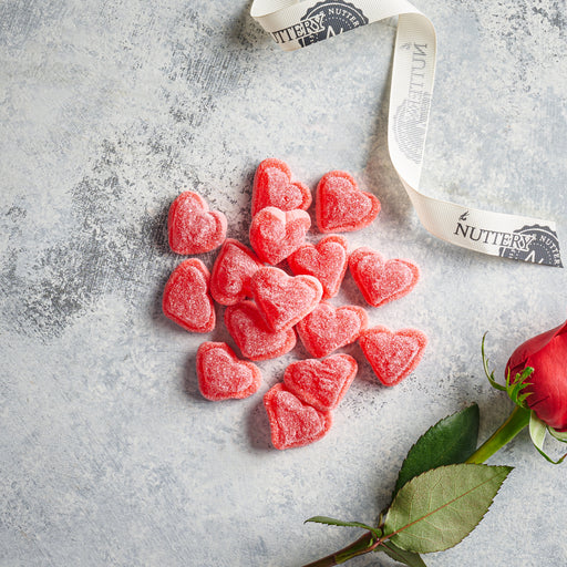 A collection of red, sugar-coated heart-shaped gummies arranged on a textured gray surface. A Nuttery-branded ribbon curls beside them, with a red rose in the corner, creating a romantic and festive Valentine's Day treat display.