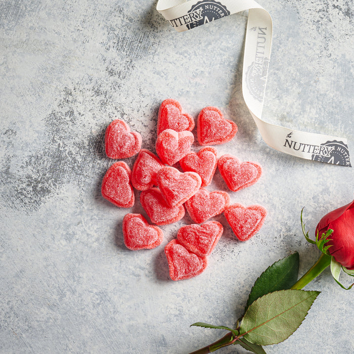A collection of red, sugar-coated heart-shaped gummies arranged on a textured gray surface. A Nuttery-branded ribbon curls beside them, with a red rose in the corner, creating a romantic and festive Valentine's Day treat display.