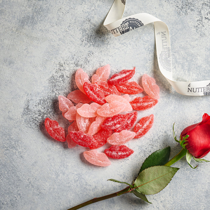 Red and pink sour lips-shaped candies sprinkled with sugar, paired with a single red rose and decorative ribbon on a textured gray surface