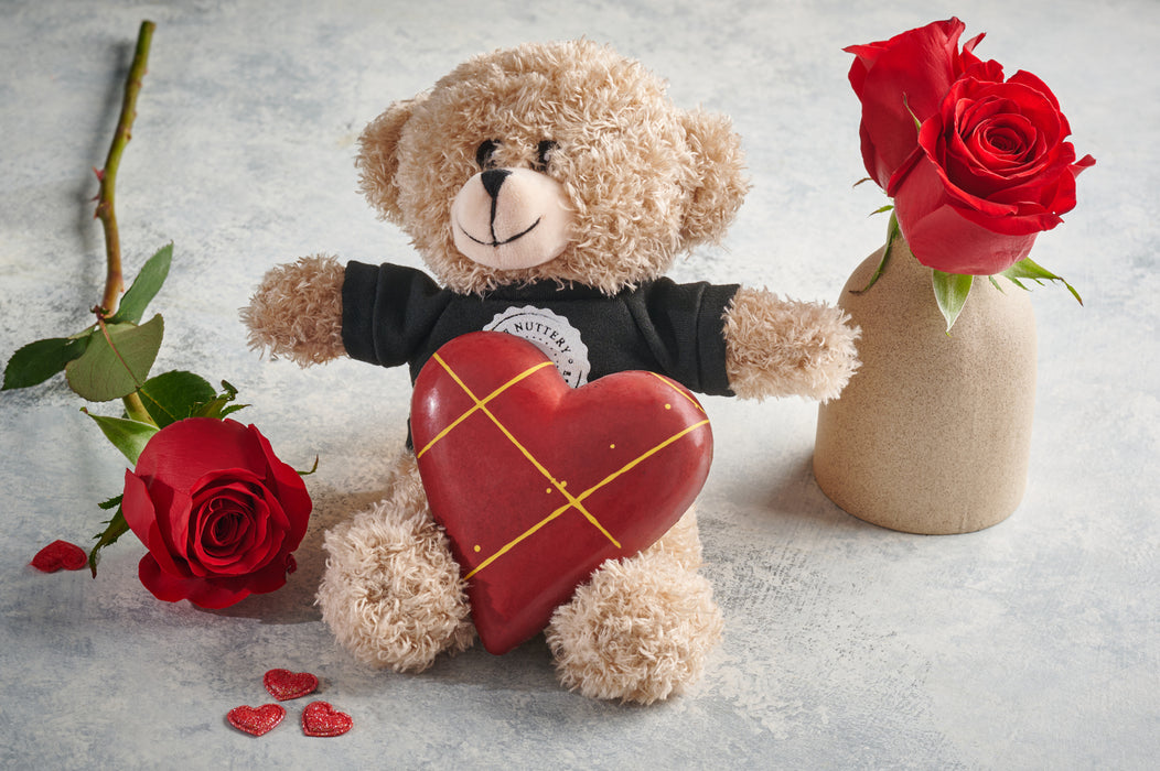 Teddy bear holding a red chocolate heart with gold accents, surrounded by roses for a romantic gift display.