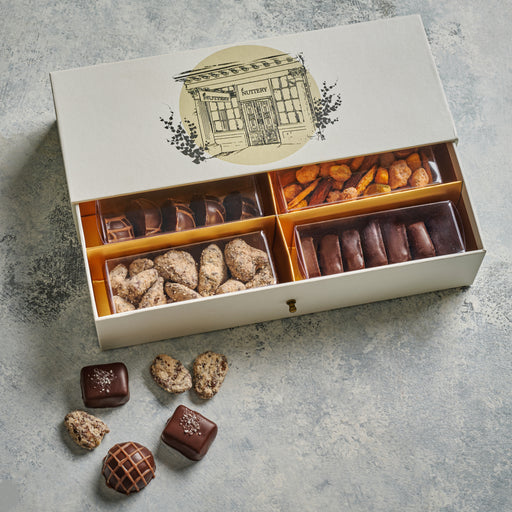 A sophisticated gift box featuring an illustration of The Nuttery storefront, filled with an assortment of gourmet chocolates, nuts, and specialty confections.