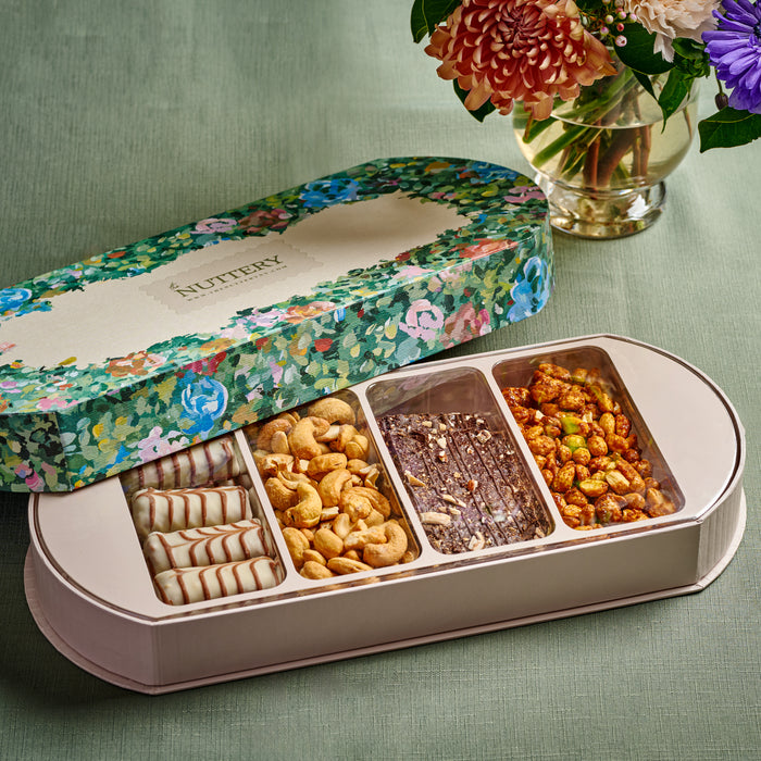 A beautifully designed floral-themed gift box from The Nuttery, opened to reveal an assortment of gourmet chocolates, roasted cashews, and glazed nuts. Set against a soft green background with elegant floral arrangements, this premium treat selection is ideal for gifting.