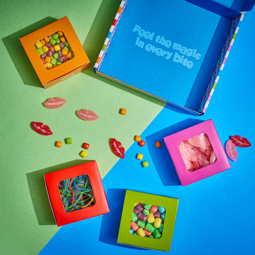 A vibrant selection of colorful gummy candies displayed in four bright boxes, arranged beside an open blue gift box with the phrase "Feel the magic in every bite." Assorted fruit-flavored sweets and gummy lips add a fun and playful touch to this festive assortment.