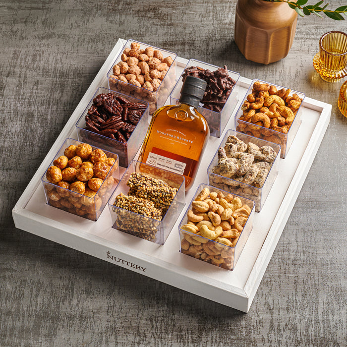 A luxurious white gift tray featuring nine compartments filled with roasted nuts, candied pecans, spiced almonds, and chocolate treats, paired with a bottle of whiskey and styled with gold accents on a gray surface