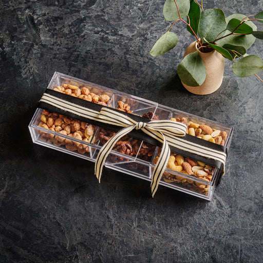 A sleek, transparent acrylic box containing a variety of gourmet nuts and chocolates, tied with a black-and-gold ribbon for an elegant presentation. Placed on a dark stone surface with a minimalist vase of greenery, this sophisticated gift set is ideal for corporate and holiday gifting.