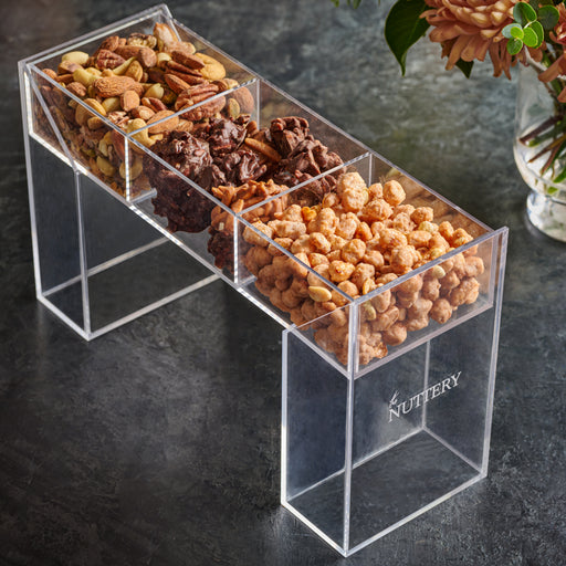 A modern acrylic stand filled with three sections of gourmet treats, including a mix of roasted nuts, chocolate clusters, and honey-glazed snacks. Displayed on a dark textured surface with a floral arrangement in the background, this elegant gift is perfect for special occasions and luxury gifting.