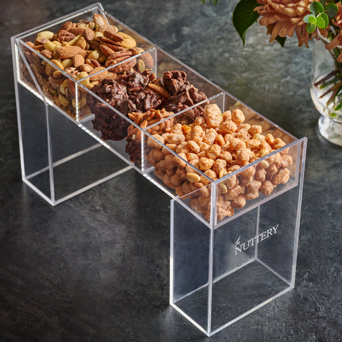 A modern acrylic stand filled with three sections of gourmet treats, including a mix of roasted nuts, chocolate clusters, and honey-glazed snacks. Displayed on a dark textured surface with a floral arrangement in the background, this elegant gift is perfect for special occasions and luxury gifting.