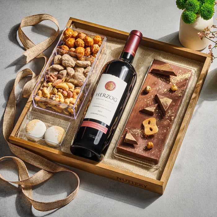 A sophisticated wine and gourmet gift set featuring a bottle of fine red wine, gourmet chocolate, and premium nuts, elegantly arranged in a wooden tray.