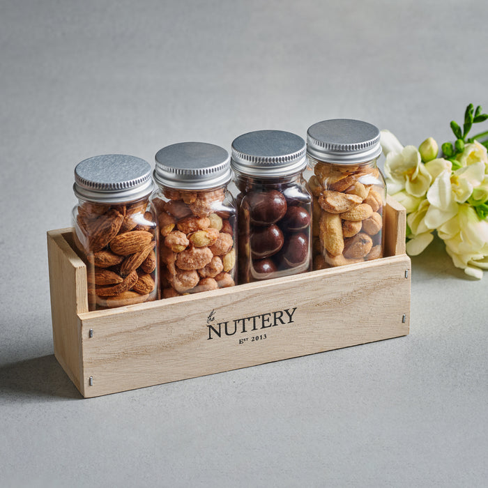 Wooden gift crate featuring four glass jars filled with almonds, candied nuts, chocolate-covered nuts, and roasted peanuts, topped with silver lids and accompanied by white floral decor, perfect for Purim gifting.