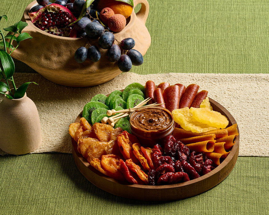 A round wooden tray featuring an assortment of dried fruits, including apricots, pineapple slices, cranberries, and kiwi, paired with hazelnut cream in the center. Styled alongside fresh grapes and pomegranates in a rustic bowl on a green tablecloth.