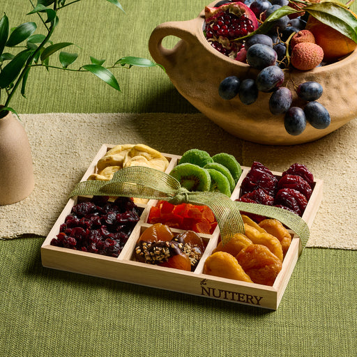 A wooden tray filled with a colorful selection of dried fruits, including kiwis, apricots, mangoes, and dates, arranged in neat rows and finished with a decorative ribbon.