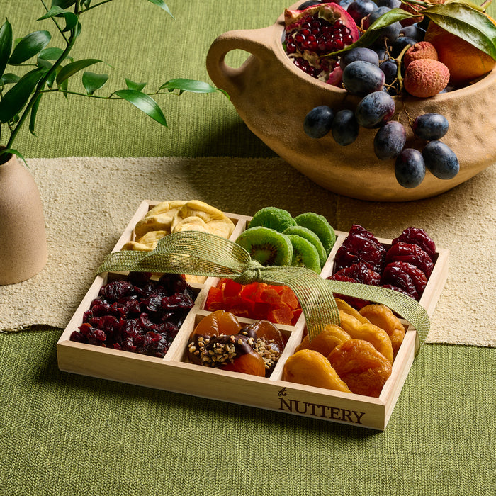 A wooden tray filled with a colorful selection of dried fruits, including kiwis, apricots, mangoes, and dates, arranged in neat rows and finished with a decorative ribbon.