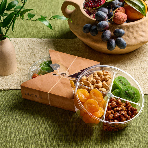 A four-section clear tray containing dried apricots, candied walnuts, roasted cashews, and dried kiwi slices, packaged in a triangular Nuttery-branded box. Styled with fresh fruits and greenery on a textured green tablecloth.
