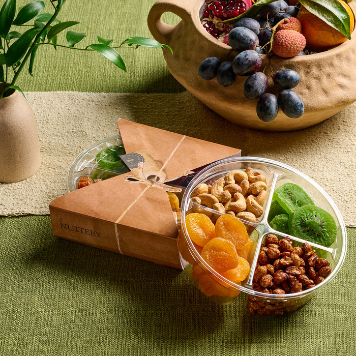 A four-section clear tray containing dried apricots, candied walnuts, roasted cashews, and dried kiwi slices, packaged in a triangular Nuttery-branded box. Styled with fresh fruits and greenery on a textured green tablecloth.