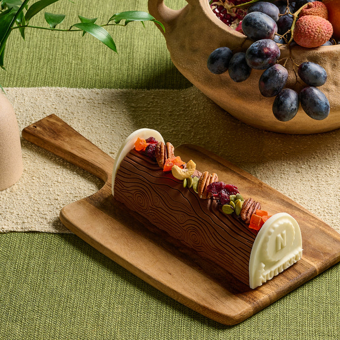 A handcrafted chocolate log from The Nuttery, designed to resemble a tree trunk, decorated with dried fruits and nuts, and finished with white chocolate end caps embossed with The Nuttery’s signature logo.