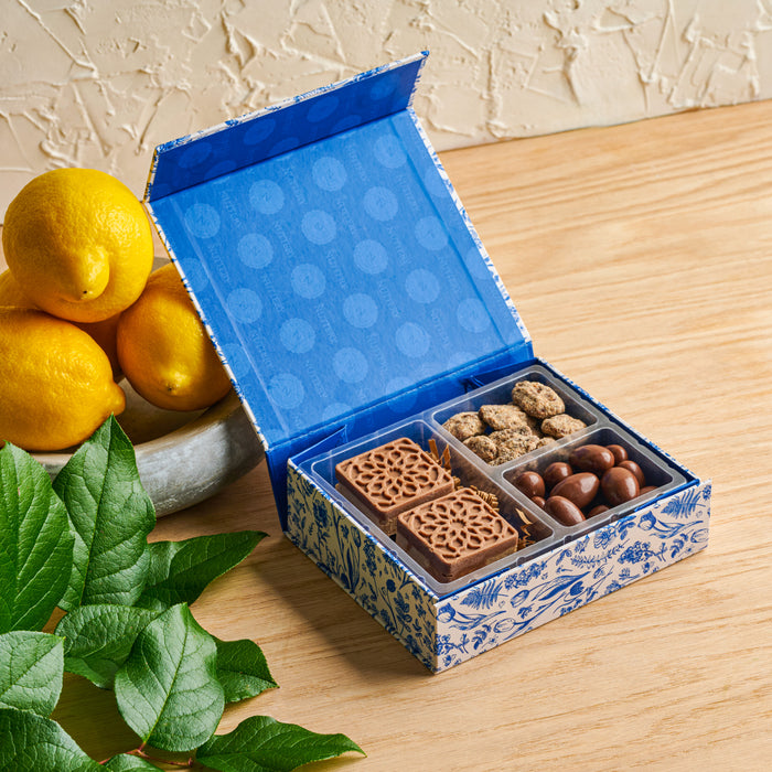 An open blue and white floral gift box from The Nuttery, revealing an assortment of chocolate-covered nuts and decorative chocolate squares.