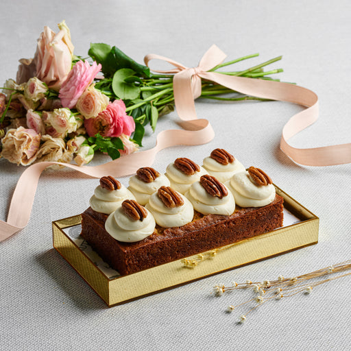 Carrot loaf topped with cream cheese frosting and pecans, elegantly displayed in a gold acrylic gift  alongside a bouquet of pastel flowers.