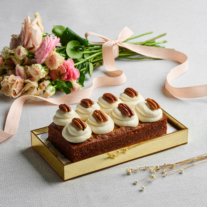 Carrot loaf topped with cream cheese frosting and pecans, elegantly displayed in a gold acrylic gift  alongside a bouquet of pastel flowers.