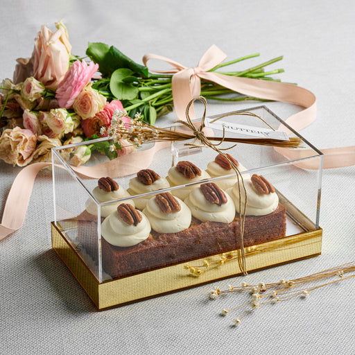 Carrot loaf topped with cream cheese frosting and pecans, elegantly displayed in a gold acrylic gift box alongside a bouquet of pastel flowers.