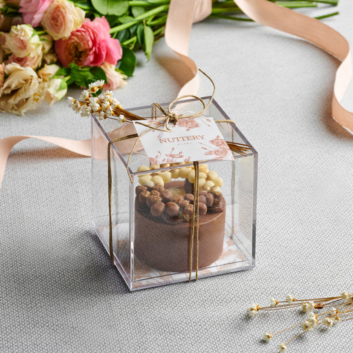 A beautifully crafted chocolate cheesecake ring, adorned with white and gold chocolate pearls, packaged in a transparent gift box with a ribbon and tag from The Nuttery.