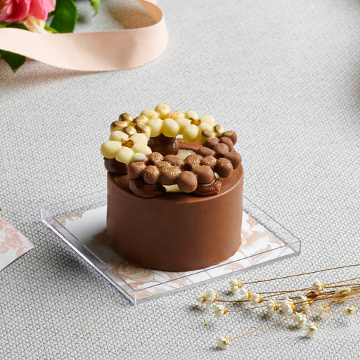 A delicate chocolate cheesecake ring decorated with edible floral elements, set on a marble tray with a soft floral arrangement in the background.