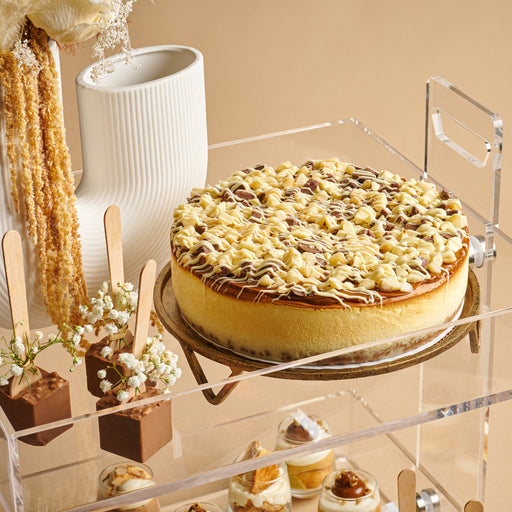 An exquisite cheesecake drizzled with white and milk chocolate sits atop a clear serving tray. Paired with delicate floral décor, this dessert setup is ideal for any festive occasion.