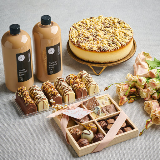 An elegant Shavuos dessert package featuring caramel and vanilla latte bottles, a round gift box tied with a cream ribbon, assorted chocolates, and nut-topped dairy desserts, paired with fresh roses on a beige background