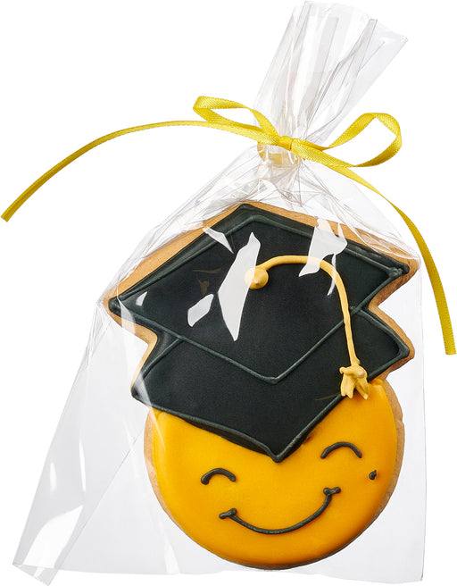 A cheerful, emoji-style graduation cookie with a black mortarboard cap and tassel, individually wrapped in a gift bag with a yellow ribbon.