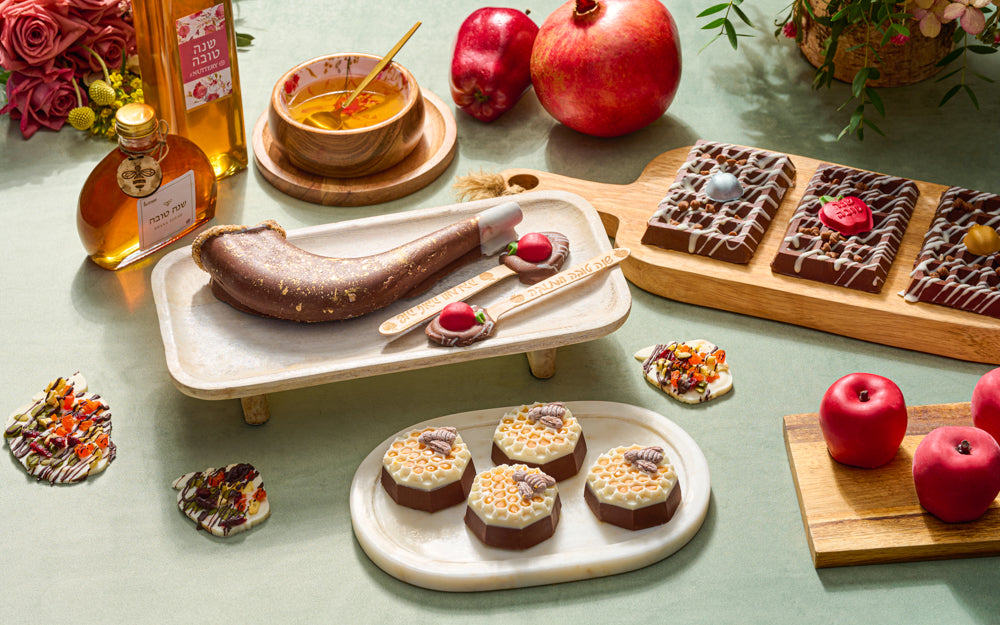 A festive Rosh Hashanah arrangement featuring a decorative chocolate shofar with gold accents, surrounded by honey jars, fresh apples and pomegranates, and gourmet chocolates on a green tablecloth