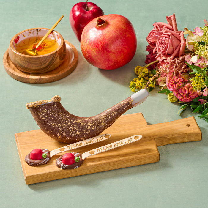 A chocolate shofar with gold detailing, paired with chocolate spoons decorated with apple designs, displayed on a wooden board alongside honey, fresh pomegranates, and a floral arrangement
