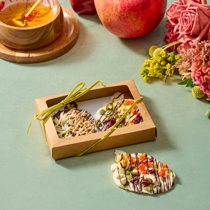 A gift box containing two apple-shaped chocolate barks topped with colorful dried fruits, nuts, and drizzled chocolate, styled alongside honey, a fresh pomegranate, and a floral arrangement on a green surface