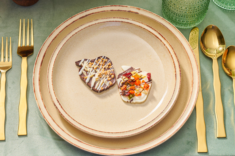 Two apple-shaped chocolate barks topped with dried fruits, nuts, and drizzled chocolate, displayed on stacked beige plates with gold cutlery and green glassware for a festive table setting