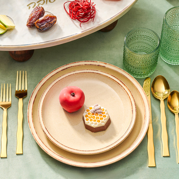 A festive Rosh Hashanah table setting featuring a honeycomb-shaped chocolate dessert and a red chocolate apple on beige plates, surrounded by gold cutlery and green glassware