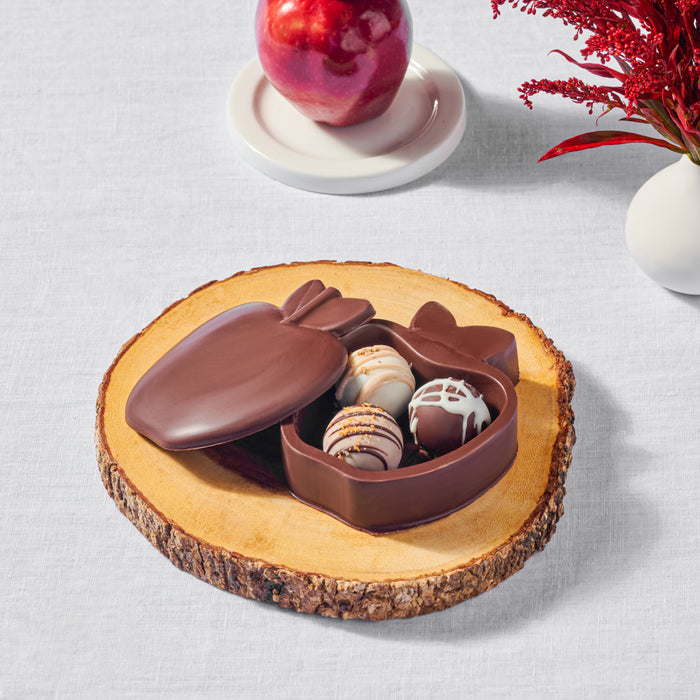 Chocolate apple-shaped box filled with gourmet truffles, displayed on a wooden slice, accompanied by a fresh apple and red floral decor, perfect for gifting or festive occasions.