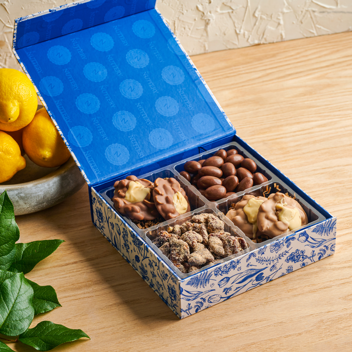 A large blue and white floral gift box from The Nuttery, open to reveal an assortment of chocolate and candied nuts.