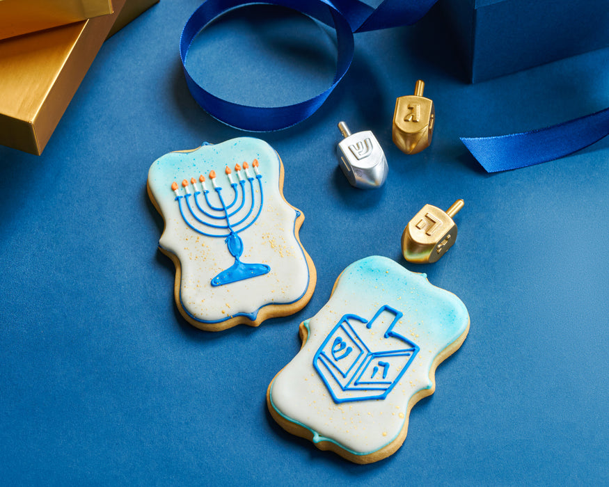 A beautifully arranged Hanukkah-themed cookie display, featuring sugar cookies decorated with a menorah and dreidel, surrounded by gold and silver dreidels and festive blue ribbons. A luxurious holiday treat, perfect for gifting or celebrations.
