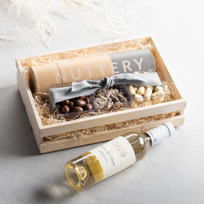 A rustic wooden gift crate filled with The Nuttery’s finest gourmet treats, including chocolates, nuts, and a bottle of white wine, wrapped with a decorative bow.