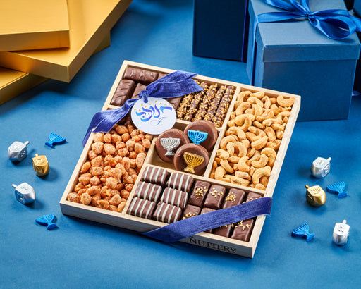 The Nuttery NY Chanukah variety gift tray, featuring a festive assortment of gourmet chocolates, roasted nuts, and menorah-decorated chocolate treats, beautifully wrapped with a blue holiday ribbon.