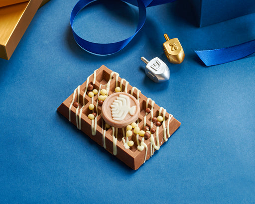 Chanukah-themed dairy waffle made of premium milk chocolate, topped with decorative white chocolate drizzle and festive sprinkles, displayed on a blue background alongside dreidels.