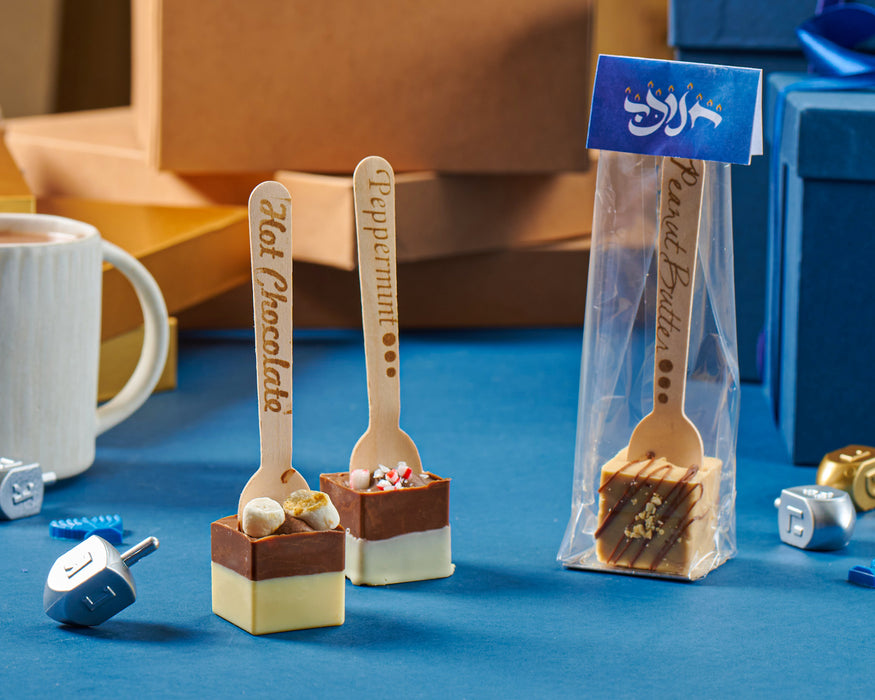 Chanukah-themed hot chocolate dippers crafted with layers of premium milk, dark, and white chocolate, topped with festive toppings like marshmallows, peppermint pieces, and caramel drizzle, displayed alongside dreidels on a blue background.