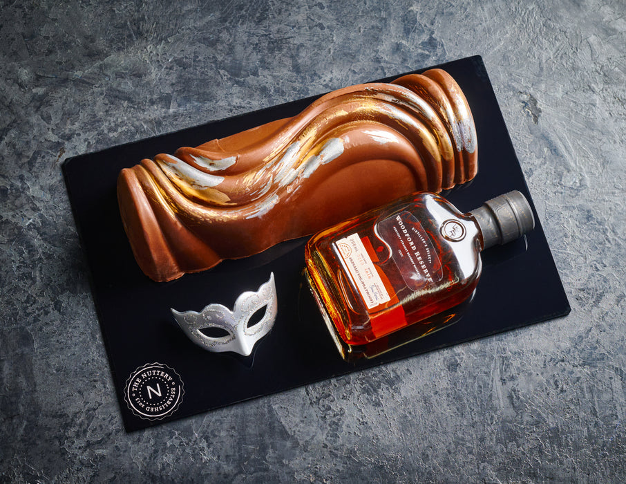 A striking chocolate swirl log from The Nuttery, featuring a glossy copper-toned exterior with silver accents, presented alongside a bottle of fine liquor and a masquerade mask for a touch of sophistication.