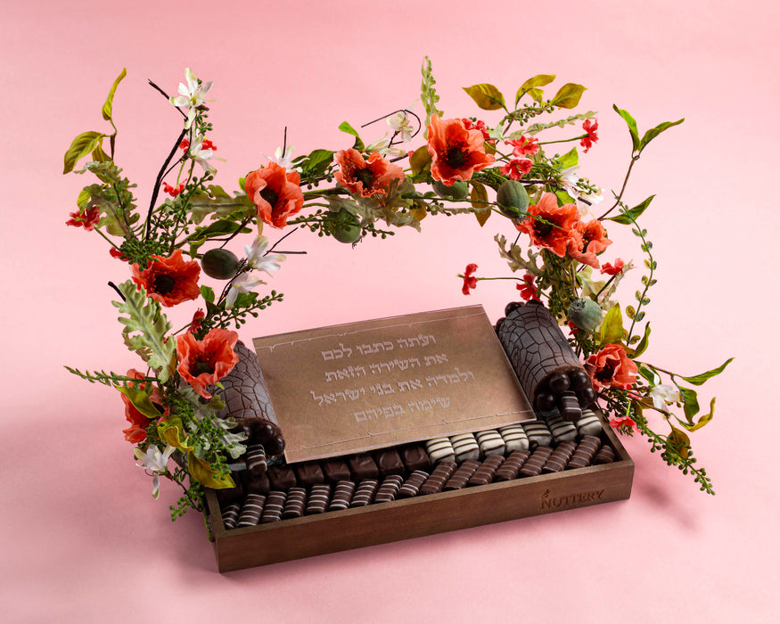 A decorative Shavuos Torah-themed display featuring a wooden tray filled with gourmet chocolates, a miniature Sefer Torah, and a floral arch of red and green blossoms, set against a soft pink background