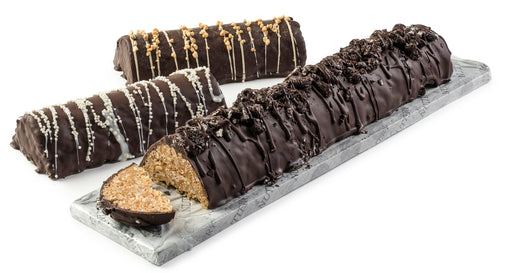 A rich peanut chew log coated in dark chocolate and topped with chocolate drizzle, displayed on a marble platter with one slice cut to reveal its crunchy peanut interior.