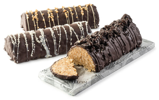 A luxurious peanut chew log coated in dark chocolate and garnished with chocolate drizzle, styled on a marble platter alongside two additional logs decorated with white and caramel drizzle.