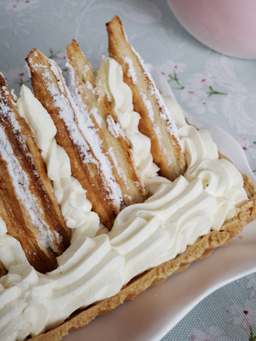 Rectangular tart topped with powdered sugar-coated cookies, displayed in a silver acrylic serving tray and tied with a branded cream ribbon, perfect for festive occasions.